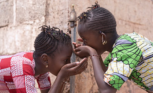 Two girls with plaited hair and wearing African-print tops drink water from a tap