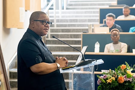 University of Pretoria Interim Vice-Chancellor and Principal Professor Themba Mosia, standing before a mike at the podium in an auditorium wearing glasses and a short-sleeved black shirt, welcoming guests to the celebration.