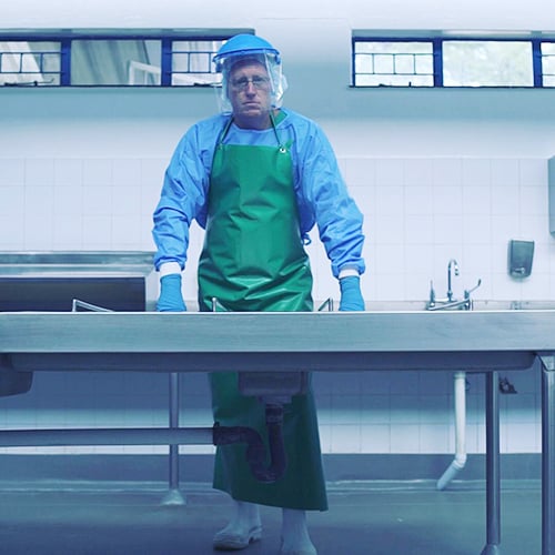 Professor Ryan Blumenthal standing behind an autopsy table dressed in blue surgical protective gear, blue surgical gloves, white rubber boots, hair net, face shield and a long green vinyl apron. Both his hands rest on the table.