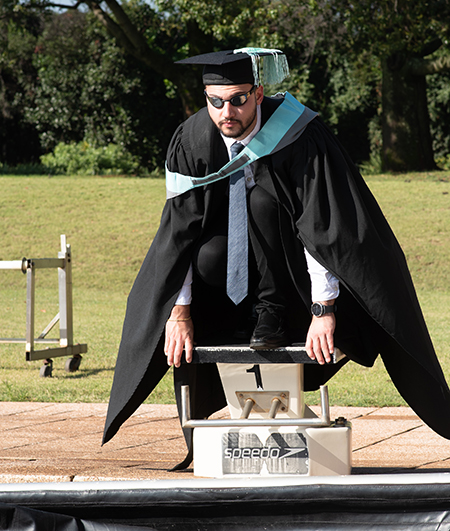 UP engineering graduate Chase Onorati perched on a swimming pool diving platform dressed in a graduation gown and cap while wearing swimming goggles on