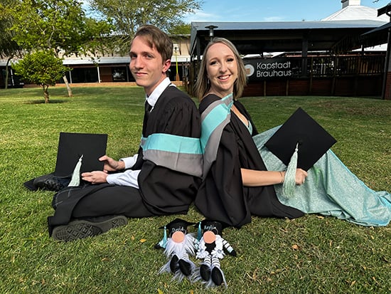 Kaylie and David sit back to back on green grass in their graduation gowns with their caps in hand while two gnomes made by their mother to represent them lie in the foreground