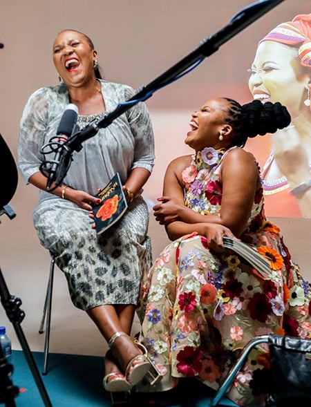Mother an daughter writers Nombeko and Sihle-isipho Nontshokweni laugh heartily as they sit in a studio with a mike infront of them. Nombeko wears a grey shimmering dress with black cheetah spots that from the bottom up. Sihle-isipho wears a colourful dress with appliqué flowers