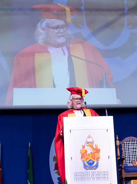 Dr Verne Sheldon Harris stands infront of a mic at a podium with a big UP logo on it. He is dressed in a red graduation gown with yellow down the front and red cap with a yellow tassel. There is a live image of him broadcasting from a big screen behind him