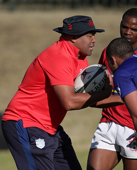 A close shot of TuksRugby new head coach Dewey Swartbooi in a red shirt, black track pants and a black hat in a training session. Partly in the shot are two players