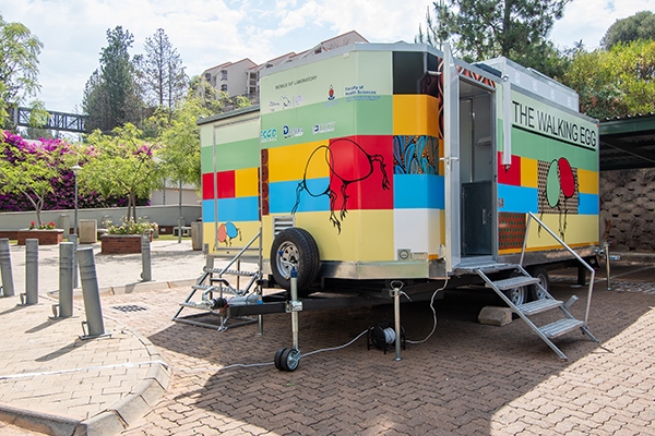 A picture of a colourful mobile lab taken from outside. It is branded in the red, yellow and blue colour blocks of the Walking Egg foundation. The organisation's name is in black on the front of the mobile unit
